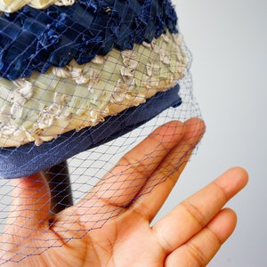 May include: A vintage hat with a blue and cream woven design, with a fine blue netting. The hat has a structured brim and a decorative pattern, showcasing a classic style.