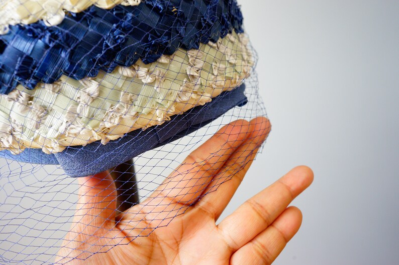 May include: A vintage hat with a blue and cream woven design, with a fine blue netting. The hat has a structured brim and a decorative pattern, showcasing a classic style.