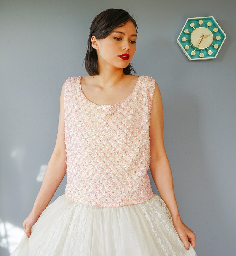 Pu&ograve; includere: Una donna indossa una maglia rosa con paillettes e una gonna di tulle bianca con bordo in pizzo. &Egrave; in piedi davanti a un muro grigio con un orologio verde acqua e bianco.