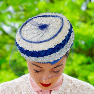 May include: A retro hat in blue and white, with a circular design and a fine blue netting veil. The hat has a woven texture and is worn by a person with red lipstick and a pink shirt.