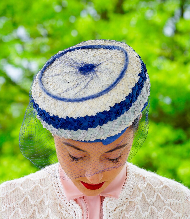 May include: A retro hat in blue and white, with a circular design and a fine blue netting veil. The hat has a woven texture and is worn by a person with red lipstick and a pink shirt.