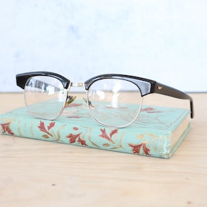 Puede incluir: Gafas con montura de plástico negro con detalles dorados y lentes transparentes que descansan sobre un pequeño libro azul verdoso con diseños florales rojos.