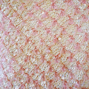Pu&ograve; includere: Primo piano di un tessuto rosa con un motivo di piccole paillettes bianche e rosa. Le paillettes sono disposte in un motivo ripetitivo di cerchi sovrapposti.