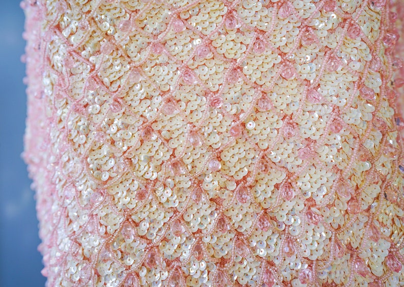 Pu&ograve; includere: Primo piano di un tessuto rosa con un motivo di piccole paillettes bianche e rosa. Le paillettes sono disposte in un motivo ripetitivo di cerchi sovrapposti.