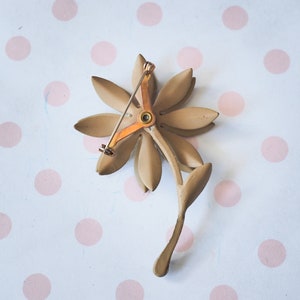 K&ouml;nnte beinhalten: Eine goldfarbene Blumenbrosche mit einem zarten Stift. Die Blume hat f&uuml;nf Bl&uuml;tenbl&auml;tter und eine Mitte mit einem kleinen, runden, goldfarbenen Akzent.