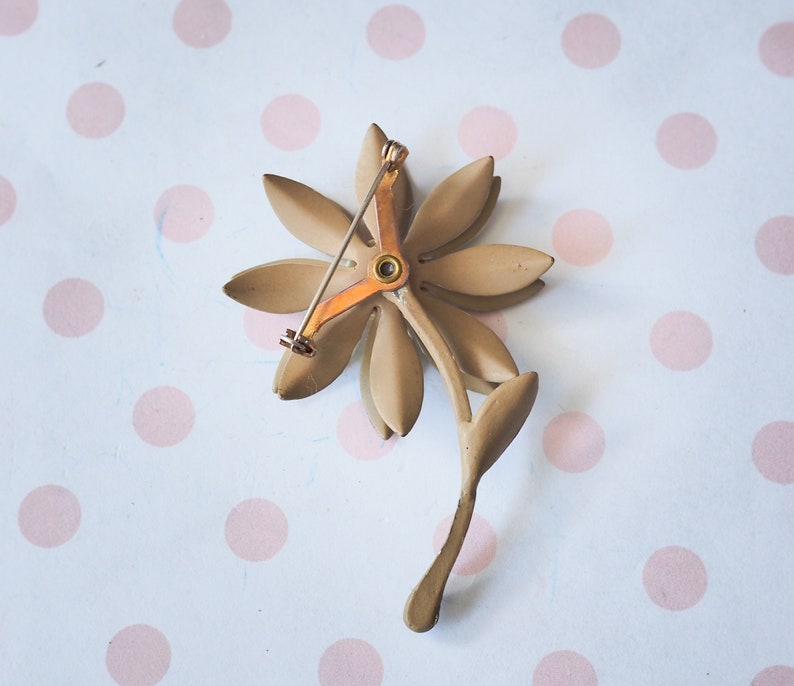 K&ouml;nnte beinhalten: Eine goldfarbene Blumenbrosche mit einem zarten Stift. Die Blume hat f&uuml;nf Bl&uuml;tenbl&auml;tter und eine Mitte mit einem kleinen, runden, goldfarbenen Akzent.