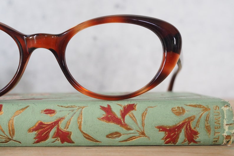 May include: A pair of tortoiseshell oval-shaped spectacles resting on a vintage book with a green cover and gold floral designs.