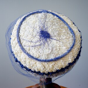 May include: A vintage hat with a white, textured crown and a blue trim. The hat features a blue netting veil and a blue circular accent. The hat is displayed on a stand, showcasing its classic design and colour scheme.