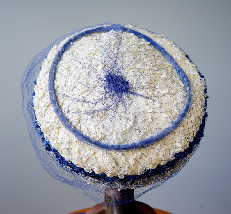 May include: A vintage hat with a white, textured crown and a blue trim. The hat features a blue netting veil and a blue circular accent. The hat is displayed on a stand, showcasing its classic design and colour scheme.