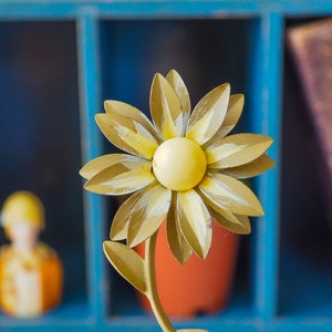K&ouml;nnte beinhalten: Eine gelbe Metallmargerite mit wei&szlig;em Zentrum, die Blume sitzt auf einem Metallstiel mit einem gr&uuml;nen Blatt. Die Blume befindet sich vor einem blauen Holzregal mit einem Buch auf der rechten Seite.