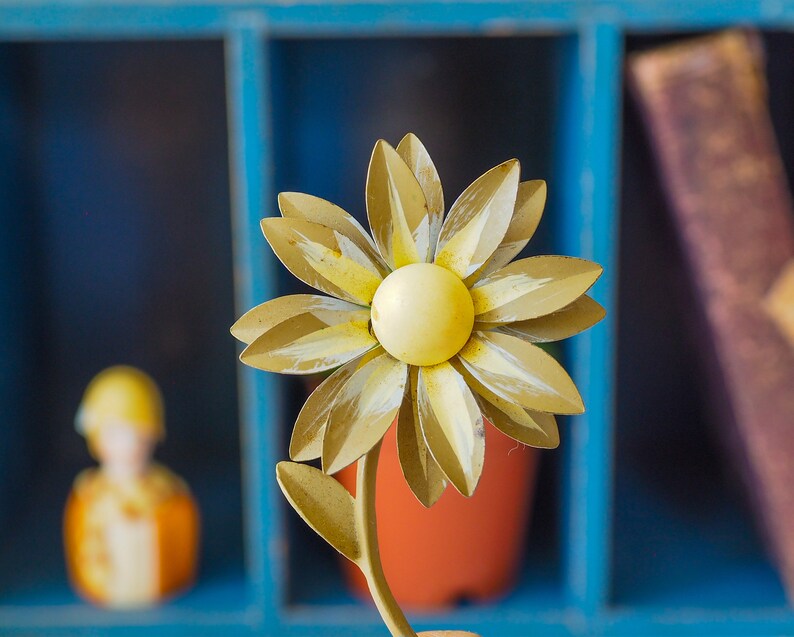 K&ouml;nnte beinhalten: Eine gelbe Metallmargerite mit wei&szlig;em Zentrum, die Blume sitzt auf einem Metallstiel mit einem gr&uuml;nen Blatt. Die Blume befindet sich vor einem blauen Holzregal mit einem Buch auf der rechten Seite.