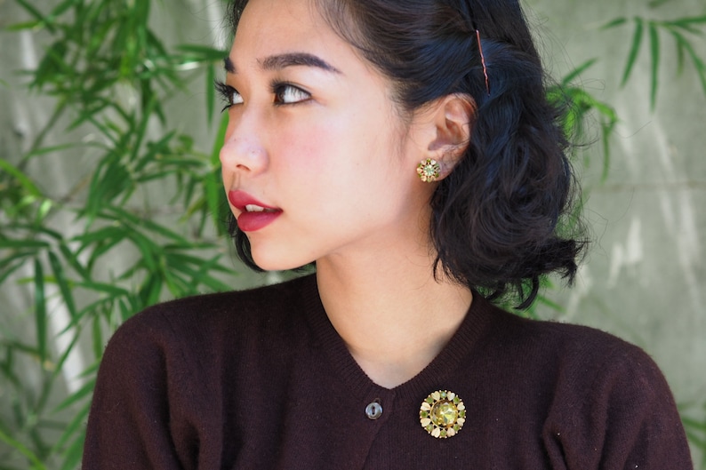 Peut inclure: Une femme aux cheveux courts et fonc&eacute;s portant un pull marron avec une broche dor&eacute;e. Elle porte des boucles d'oreilles dor&eacute;es avec des pierres vertes.