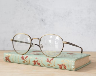 Peut inclure: Lunettes rondes à monture en métal marron avec des verres dorés, posées sur un livre vintage à couverture verte et motif floral.