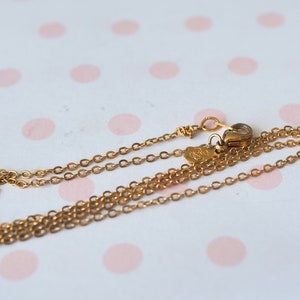 May include: A delicate gold-colored necklace with a fine chain and a small clasp. The necklace is displayed on a white surface with pink polka dots. The chain is thin and appears to be made of metal.