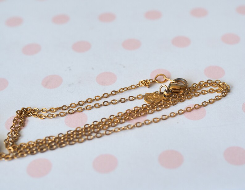 May include: A delicate gold-colored necklace with a fine chain and a small clasp. The necklace is displayed on a white surface with pink polka dots. The chain is thin and appears to be made of metal.