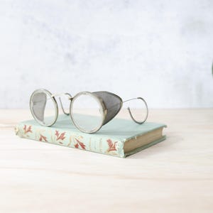 May include: Antique silver-framed spectacles with round lenses and a side shield rest on a vintage book with a faded green cover and floral design. The book is on a light wooden surface.