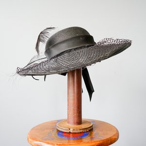 May include: A black wide-brimmed hat with a black feather and a black ribbon. The hat is on a wooden stand.