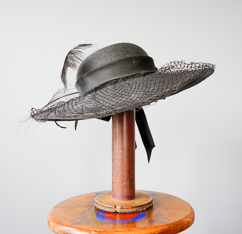 May include: A black wide-brimmed hat with a black feather and a black ribbon. The hat is on a wooden stand.
