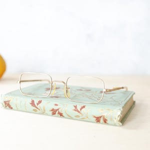 Puede incluir: Una taza de cerámica blanca con un asa negra y el texto "But First, Coffee" en negrita negra. La taza se encuentra sobre un fondo borroso, sugiriendo un ambiente acogedor.