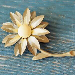 K&ouml;nnte beinhalten: Eine goldene Brosche aus Metall in Form einer Blume mit einem wei&szlig;en Zentrum und einem Stiel. Die Blume hat eine strukturierte Oberfl&auml;che und ist im Vintage-Stil.