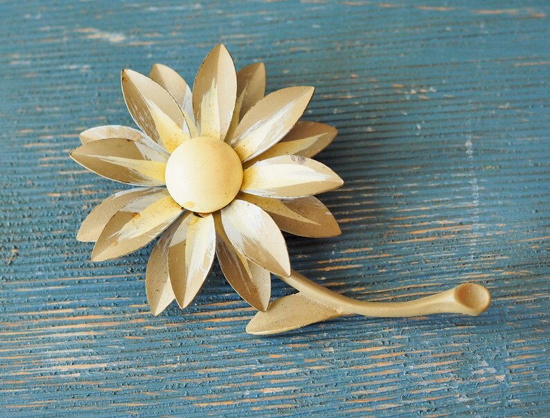 K&ouml;nnte beinhalten: Eine goldene Brosche aus Metall in Form einer Blume mit einem wei&szlig;en Zentrum und einem Stiel. Die Blume hat eine strukturierte Oberfl&auml;che und ist im Vintage-Stil.