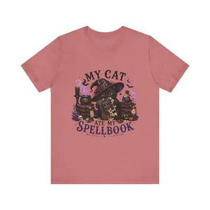 May include: A dusty rose t-shirt featuring a graphic of a black cat wearing a witch hat, surrounded by books, a cauldron, and potions. The text reads MY CAT ATE MY SPELLBOOK. The design includes purple accents, stars, and a small skull.