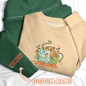 Puede incluir: Una sudadera beige con un personaje Bulbasaur bordado en verde claro con una calabaza en la espalda, rodeado de hojas de otoño. Se ve una manga de sudadera con capucha verde oscuro con el texto naranja SPOOKY TRAINER CUSTOM NAME.