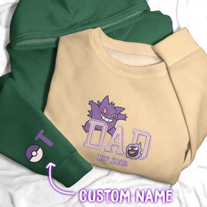 May include: A beige crewneck sweatshirt featuring a purple Gengar character graphic above the text DAD EST. 2026, layered over a dark green hooded sweatshirt with a purple letter T and a small circular purple and white ball icon on the sleeve.
