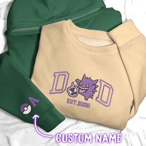 May include: A beige crewneck sweatshirt featuring an embroidered purple Gengar and Gastly graphic with the text DAD EST. 2026, layered over a dark green hoodie with an embroidered purple letter A and a Poké Ball icon on the sleeve cuff.