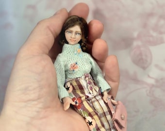 May include: A miniature doll with brown hair, wearing a light green long-sleeved sweater with small floral embroidery and plaid trousers featuring a red, white, and brown pattern. The doll carries a small pink handbag with a gold-colored snap.