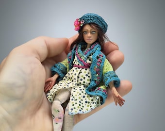 May include: A small doll with brown hair wearing a teal crocheted beret with a pink floral accent, a light green and blue crocheted cardigan, and a white dress with green and blue polka dots. The doll wears white patterned tights and brown shoes.