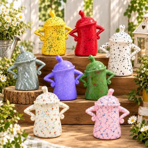 May include: A collection of eight ceramic canisters with lids, each featuring two handles positioned like arms on hips. The canisters come in various colors including yellow, red, white, grey, purple, green, and pink, with speckled or splatter paint patterns.
