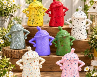 May include: A collection of eight ceramic canisters with lids, each featuring two handles positioned like arms on hips. The canisters come in various colours including yellow, red, white, grey, purple, green, and pink, with speckled or splatter paint patterns.