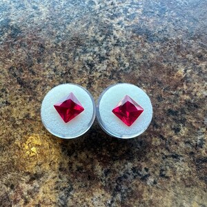 May include: Two square-cut, vibrant red gemstones are each displayed individually within a small, round, clear plastic container lined with a white foam insert. The faceted stones exhibit a sharp, pointed shape and a deep, saturated crimson color.