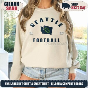 May include: A sand-coloured crewneck sweatshirt featuring dark blue text that reads SEATTLE FOOTBALL. The design includes a dark blue silhouette of the state of Washington with a green football graphic in the centre, flanked by EST. and 1976.