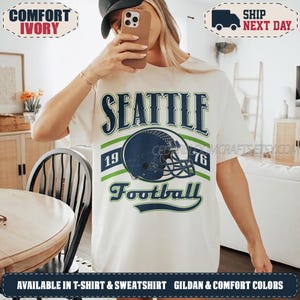 May include: An ivory t-shirt featuring a graphic print with the text SEATTLE, 1976, and Football. The design includes a dark blue football helmet centred between two horizontal green stripes, all displayed on the front of the garment.