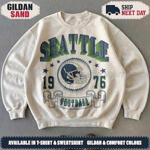 May include: A sand-colored crewneck sweatshirt featuring a graphic print with the text SEATTLE in large dark blue and green letters. Below, a football helmet illustration is centered between the numbers 19 and 76, with a banner reading FOOTBALL.