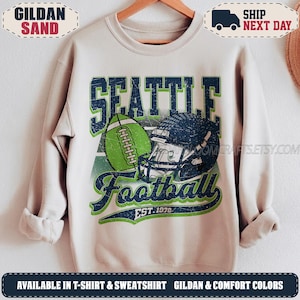 May include: A sand-colored crewneck sweatshirt featuring a distressed graphic print. The design includes the text SEATTLE in dark blue, a green football, a dark blue football helmet, and the text Football EST. 1976 in dark blue and green.