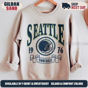 May include: A sand-colored crewneck sweatshirt featuring a dark blue and green graphic print. The design includes the text SEATTLE, the numbers 19 and 76, a football helmet illustration inside a circular border, and a banner with the word FOOTBALL.