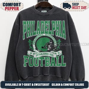Könnte beinhalten: Ein anthrazitfarbenes Sweatshirt mit Rundhalsausschnitt und einem grün-weißen Grafikdruck. Das Design zeigt den Schriftzug PHILADELPHIA FOOTBALL, eine Abbildung eines grünen Football-Helms und den Text ESTABLISHED 1933 auf einem Banner.