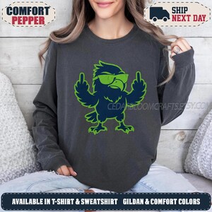 May include: A charcoal grey long-sleeve sweatshirt featuring a graphic of a dark blue eagle wearing bright green sunglasses and giving the middle finger with both wings. The eagle has a distressed, textured green outline.