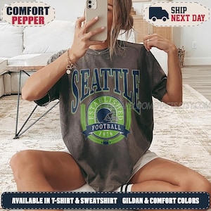 May include: A charcoal gray short-sleeve t-shirt featuring a graphic print with the text SEATTLE in large blue letters above a green and blue circular emblem containing a football helmet, the words ESTABLISHED 1976, and the word FOOTBALL.