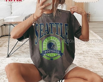 May include: A charcoal grey short-sleeve t-shirt featuring a graphic print with the text SEATTLE in large blue letters above a green and blue circular emblem containing a football helmet, the words ESTABLISHED 1976, and the word FOOTBALL.