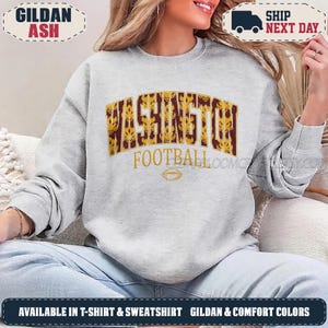May include: A light grey crewneck sweatshirt featuring the word WASHINGTON in large, gold and maroon patterned block letters, with the word FOOTBALL and a small football graphic printed in gold underneath the main text on the front.