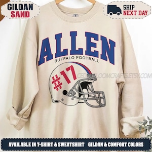 May include: A sand-coloured crewneck sweatshirt featuring the blue text ALLEN and BUFFALO FOOTBALL above a graphic of a white football helmet with a red number 17 on the side. The helmet has a grey face mask and red and blue stripe details.