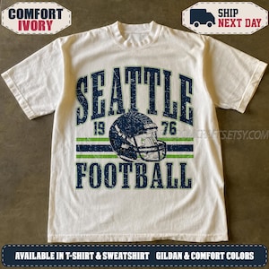 May include: An ivory short-sleeve t-shirt featuring a distressed dark blue and green graphic. The design includes the text SEATTLE 1976 and FOOTBALL, centred around a dark blue football helmet illustration with a green stripe and face mask.