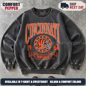 May include: A charcoal grey crewneck sweatshirt featuring a large orange graphic on the chest. The design includes the text CINCINNATI, the numbers 19 and 68, a football helmet icon inside a star-bordered circle, and a banner reading FOOTBALL.