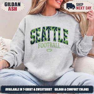 May include: A light grey crewneck sweatshirt featuring the text SEATTLE in large dark blue block letters with a green eagle pattern inside. Below, the word FOOTBALL appears in green, followed by a small green football graphic centred.