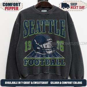 May include: A charcoal gray crewneck sweatshirt featuring a graphic print with the text SEATTLE in dark blue and green, a football helmet, the numbers 19 and 76, and the word FOOTBALL. The design includes a football field illustration.
