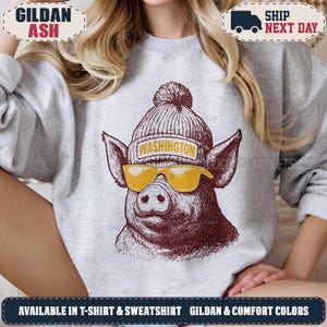 May include: A light grey crewneck sweatshirt featuring a graphic of a pig wearing a knit beanie with a pom-pom and yellow sunglasses. The beanie has the word WASHINGTON printed on a yellow band. The pig illustration is rendered in dark maroon ink.